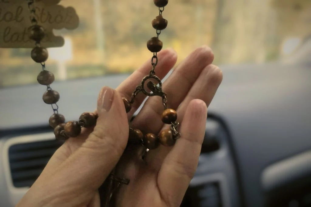Różaniec Ręka trzymająca różaniec z brązowych paciorków na tle wnętrza samochodu, z widoczną deską rozdzielczą i rozmytym tłem za szybą.