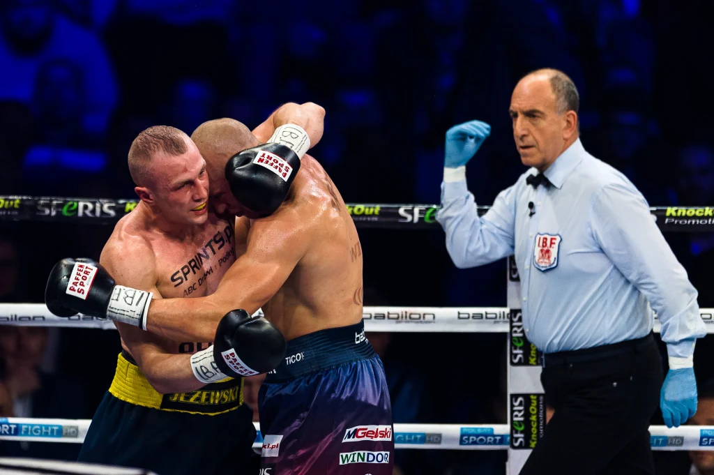 Nikodem Jeżewski (z lewej) Dwóch bokserów w ringu objętych podczas walki, arbiter uważnie obserwuje sytuację, mając uniesioną rękę gotową do reakcji. Scena oświetlona reflektorami, widoczna dynamiczna atmosfera sportowego pojedynku.