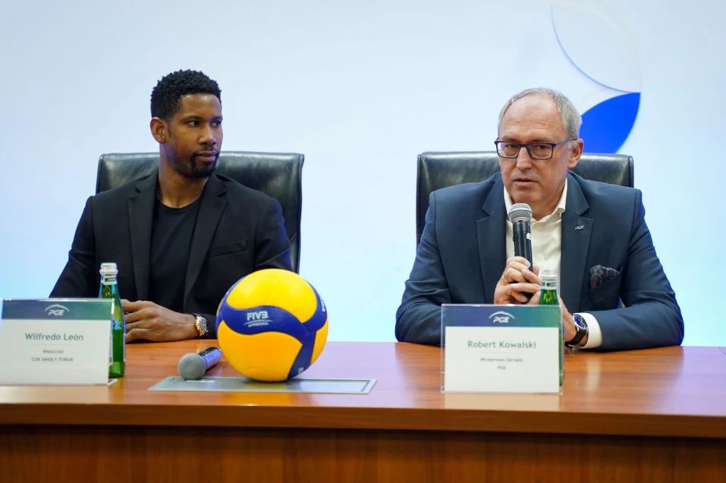Dwóch mężczyzn siedzących przy stole konferencyjnym, jeden z nich mówi do mikrofonu, na stole leży piłka do siatkówki oraz stoją dwie tabliczki z imionami, butelki z wodą i notatki.