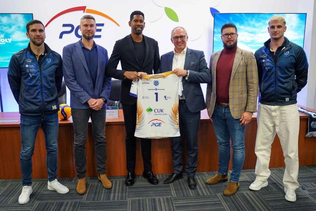 Grupa sześciu mężczyzn stoi w pomieszczeniu konferencyjnym, z czego dwie osoby w centrum trzymają koszulkę sportową z numerem 1 oraz logotypem PGE. W tle widoczne są duże monitory, elementy graficzne oraz logo sponsora.