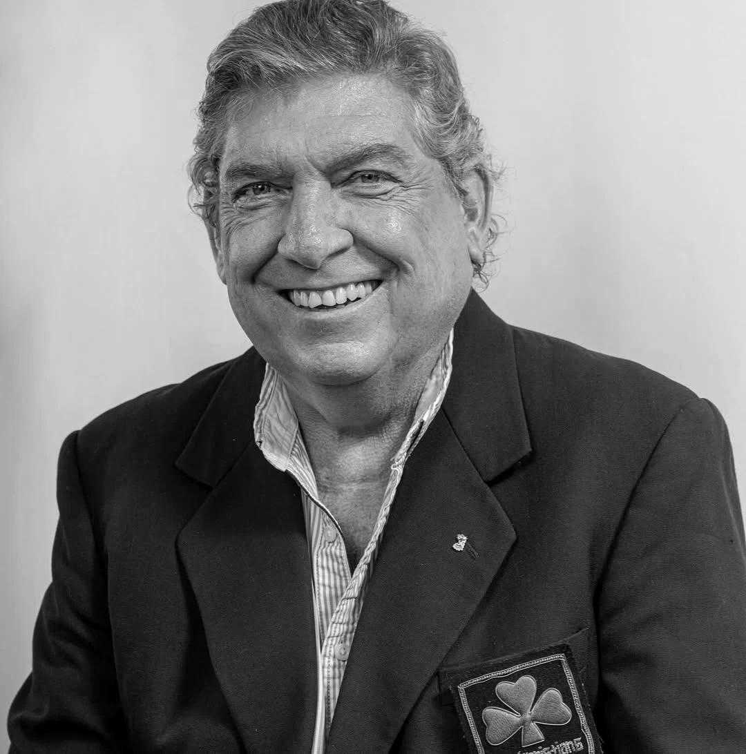 Starszy mężczyzna o siwych włosach uśmiecha się, ubrany w marynarkę z naszywką z motywem czterolistnej koniczyny, jasną koszulę i siedzi na jasnym tle.