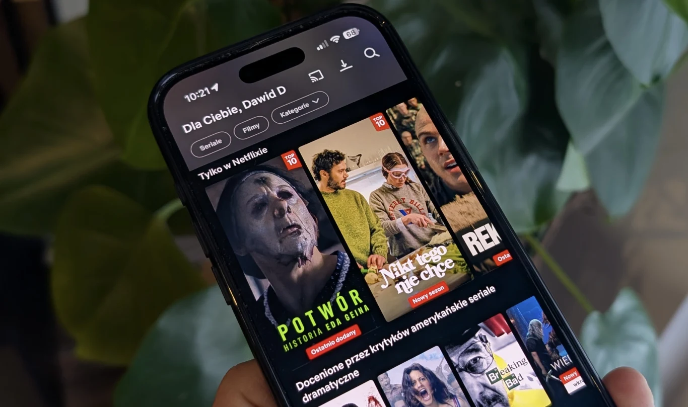 Używasz aplikacji Netfliksa? Przygotowano trzy nowe funkcje. Ekran smartfona z włączoną aplikacją z filmami i serialami, na pierwszym planie widoczne miniatury filmów, w tym horror z przerażającą postacią oraz kolorowa miniatura serialu komediowego, w tle rozmyte zielone liście rośliny.