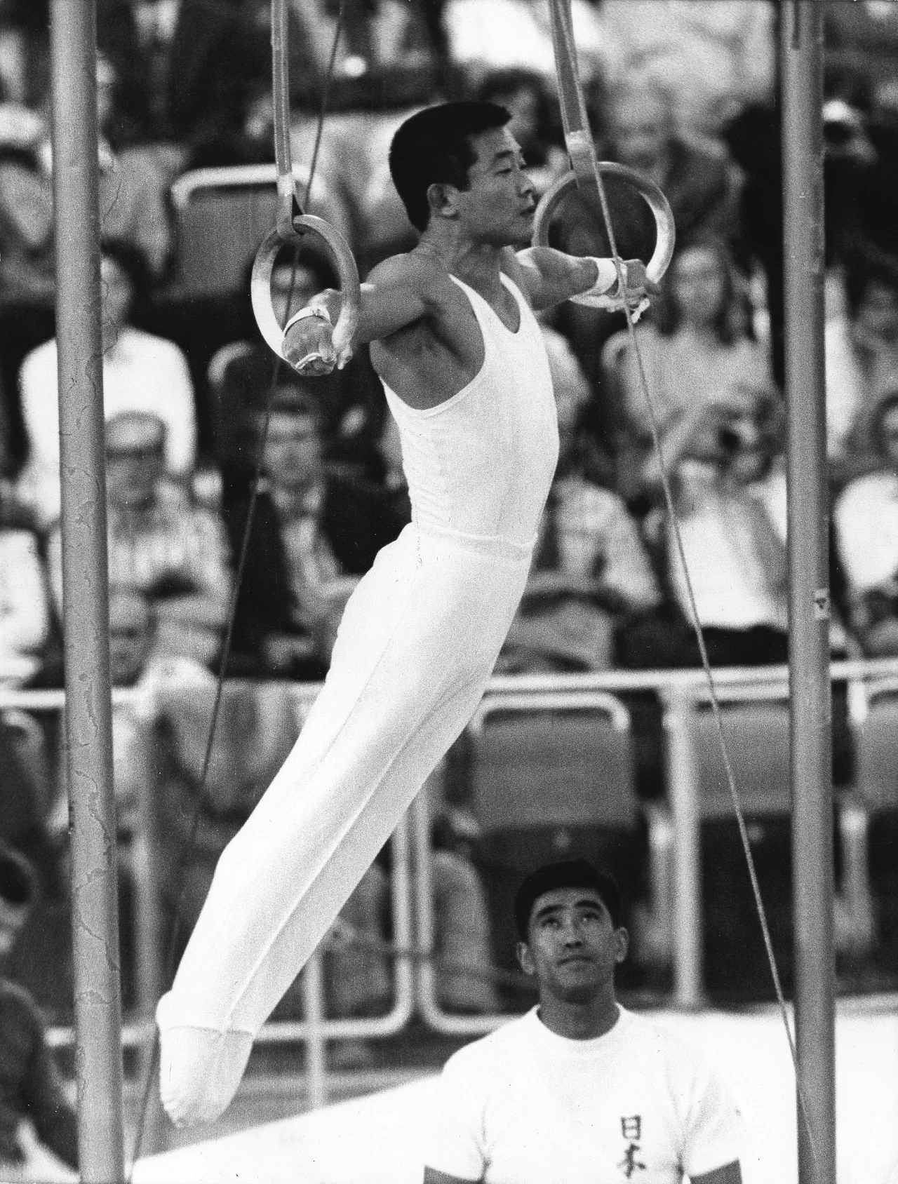 Gimnastyk w białym stroju wykonuje ćwiczenie na kółkach gimnastycznych, unosząc się nad ziemią z wyprostowanymi ramionami. W tle widoczna jest publiczność siedząca na trybunach, a poniżej gimnastyka stoi trener lub sędzia ubrany w jasny strój z napisem...