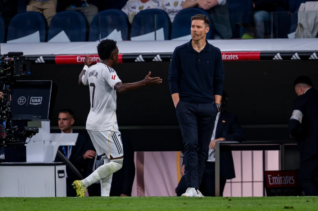 Vinicius Jr. i Xabi Alonso Piłkarz w białym stroju rozmawia żywiołowo z trenerem stojącym przy linii bocznej boiska, w tle widać ławkę rezerwowych i elementy stadionu.