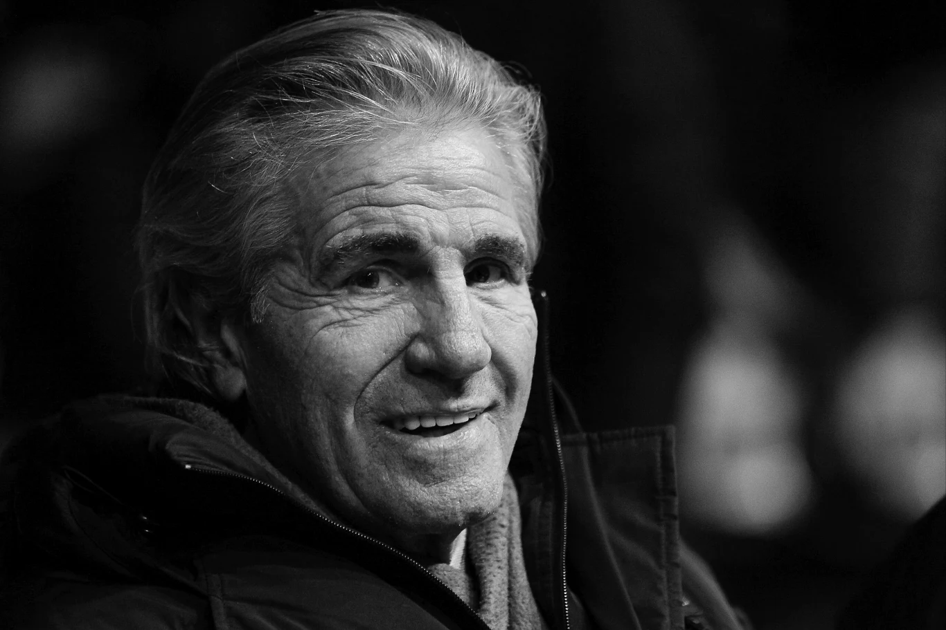 Starszy mężczyzna z siwymi włosami, uśmiechnięty, ubrany w grubą kurtkę, patrzy w stronę obiektywu na ciemnym tle.