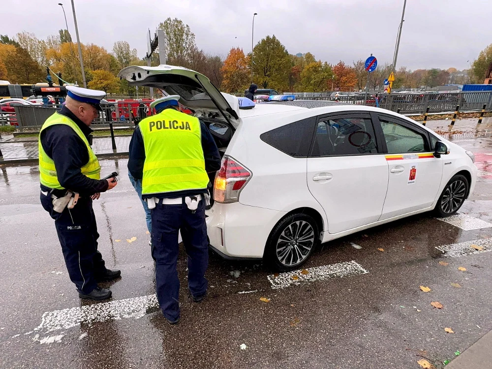 Dwóch policjantów w odblaskowych kamizelkach przeprowadza kontrolę przy białym samochodzie z otwartym bagażnikiem na parkingu, wokół jesienne drzewa i mokra nawierzchnia po deszczu.