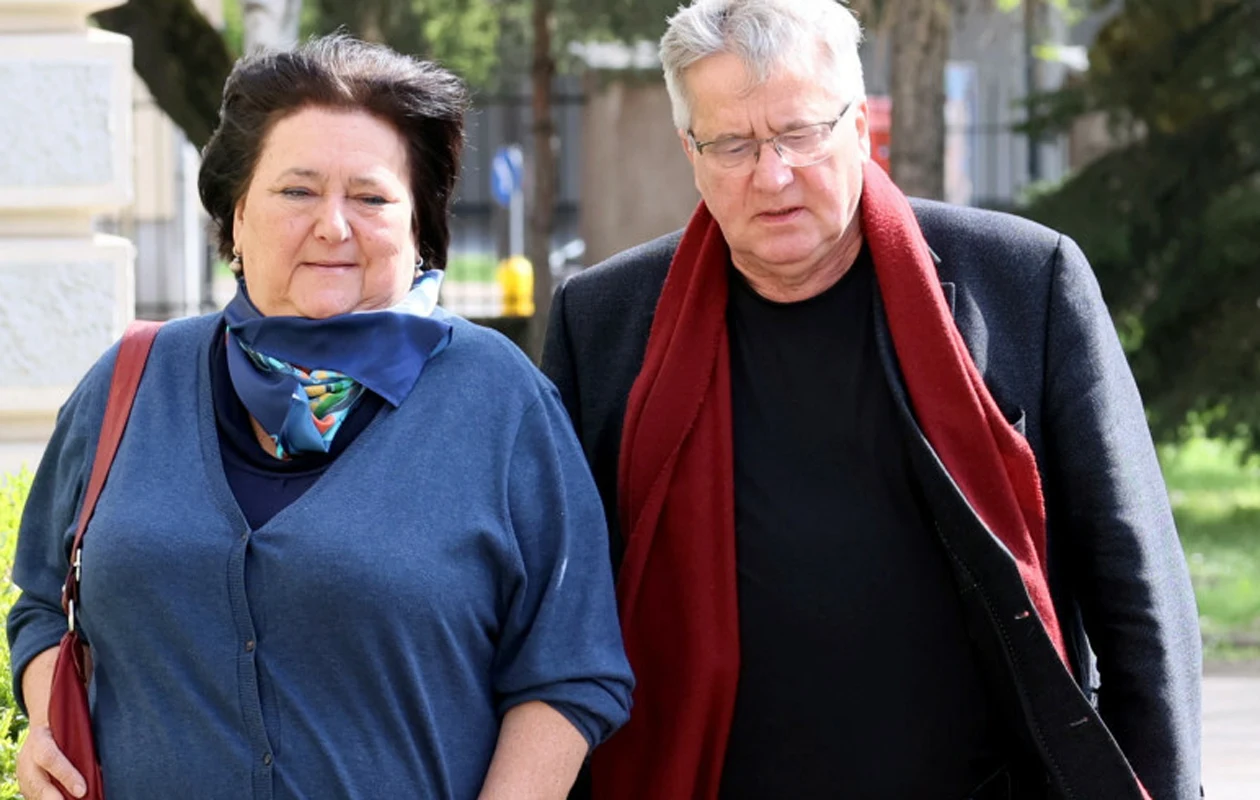 Anna i Bronisław Komorowscy Starsza kobieta i mężczyzna idą razem, ubrani w eleganckie ubrania, spacerują w słoneczny dzień w otoczeniu zieleni i budynków.