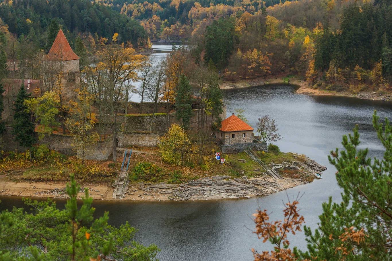 Zamek na skraju jeziora otoczony kolorowym, jesiennym lasem. Przylegająca do zamku kamienna wieża oraz budynek z czerwoną dachówką. Spokojna tafla wody otacza półwysep, a drzewa w odcieniach żółci, czerwieni i zieleni tworzą malowniczy krajobraz.