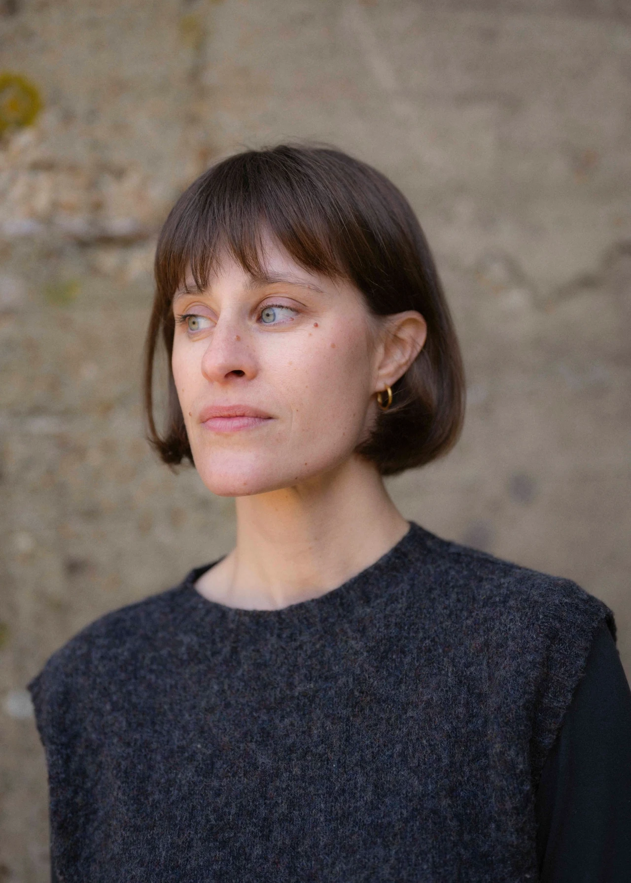 Kobieta o jasnej cerze i prostych, ciemnobrązowych włosach sięgających do brody, ubrana w ciemny, wełniany bezrękawnik i czarną bluzkę, stojąca na tle surowej, jasnej ściany z widocznymi śladami upływu czasu.