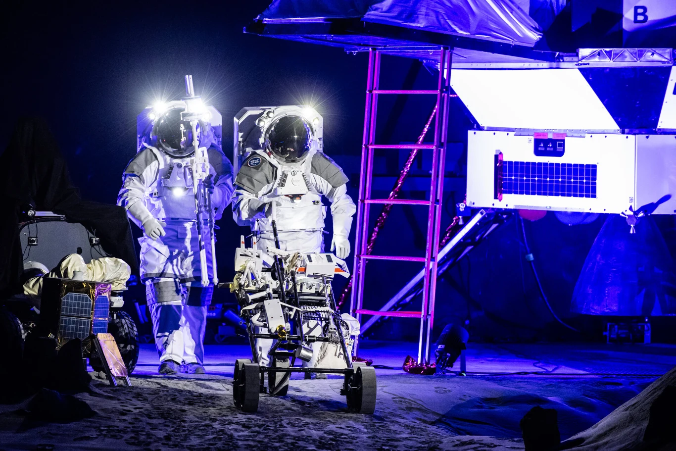 Dwóch astronautów w skafandrach kosmicznych stoi obok łazika badawczego na tle modułu kosmicznego oświetlonego światłami.