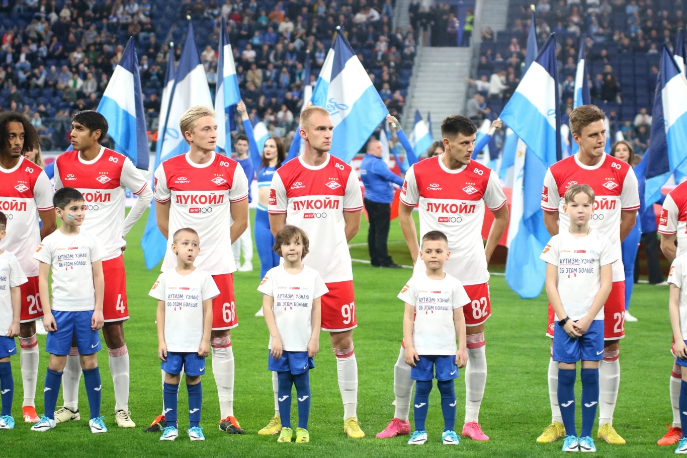 Piłkarze w czerwono-białych strojach oraz dzieci stoją w szeregu na murawie stadionu, za nimi widoczne flagi z biało-niebieskimi barwami oraz rozległa widownia wypełniona kibicami.