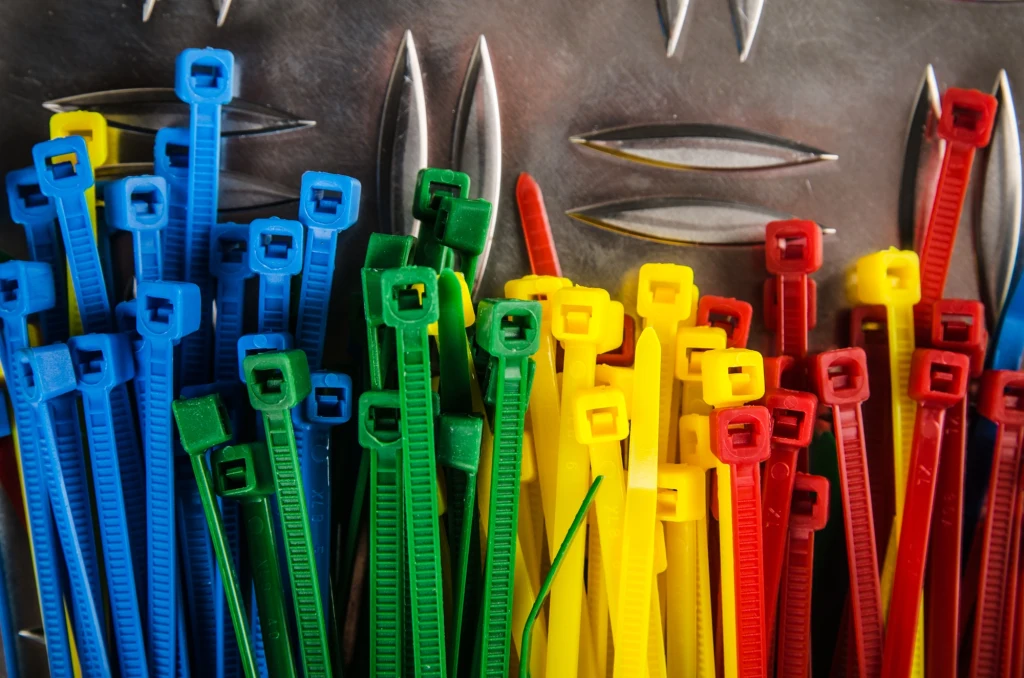 Kolorowe opaski zaciskowe w odcieniach niebieskim, zielonym, czerwonym i żółtym poukładane rzędami na tle metalowej powierzchni z otworami służącymi do ich montażu lub przechowywania.