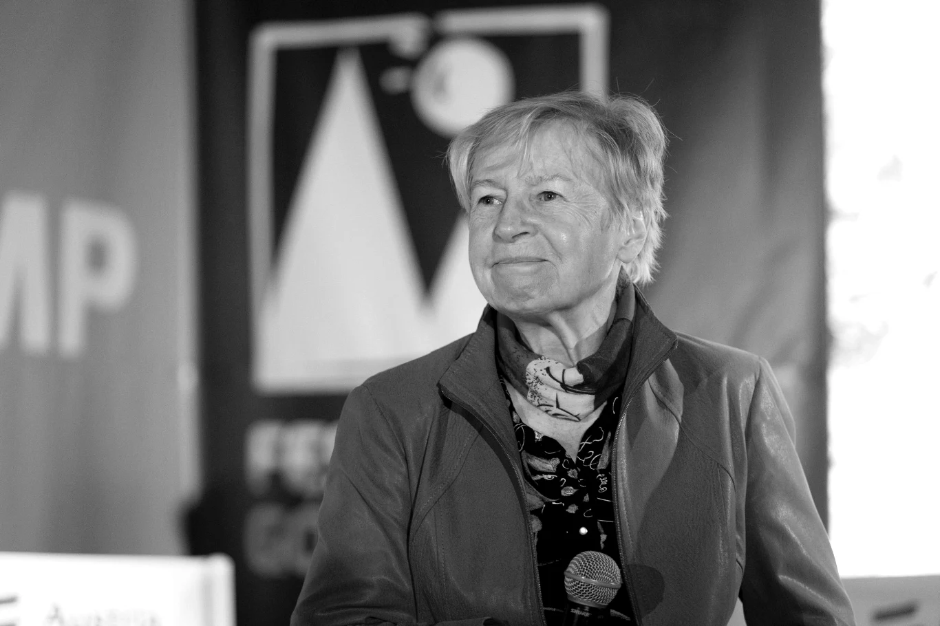 Starsza kobieta z krótko przyciętymi włosami w kurtce i apaszce, trzymająca mikrofon, stoi na tle nieostrego baneru z wyraźnym wzorem geometrycznym.