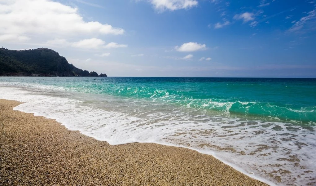 Piaszczysta plaża z delikatnie spienionymi falami, przejrzystą wodą w odcieniach turkusu oraz skalistym wzgórzem porośniętym roślinnością w tle. Na niebie kilka białych chmur, dominują błękitne barwy.