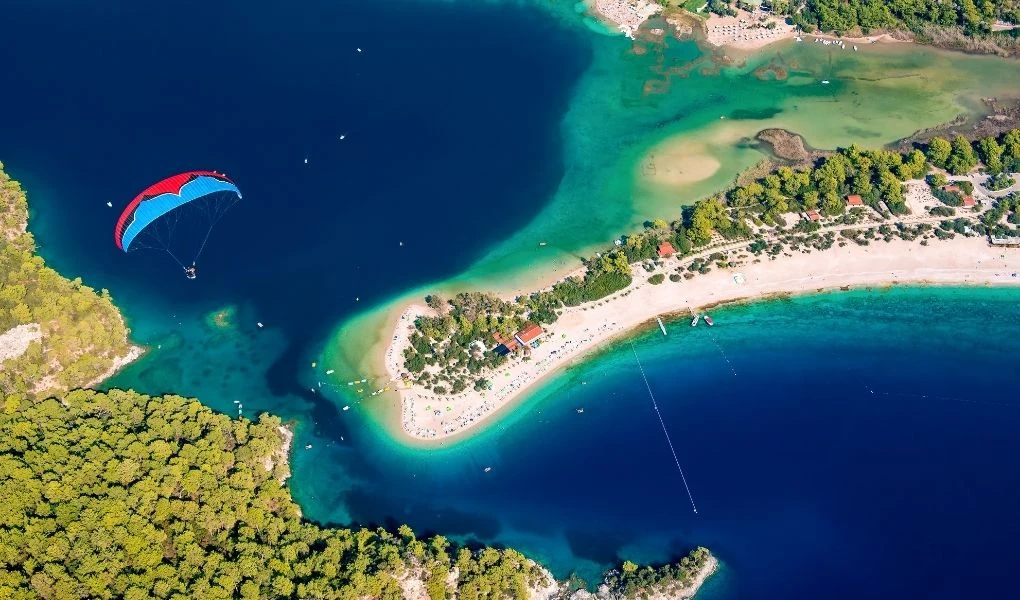Widok z lotu ptaka na półwysep pokryty lasem i piaszczystą plażą otoczoną turkusową wodą, na której można dostrzec ludzi kąpiących się i wypoczywających oraz paralotniarza nad wodą.