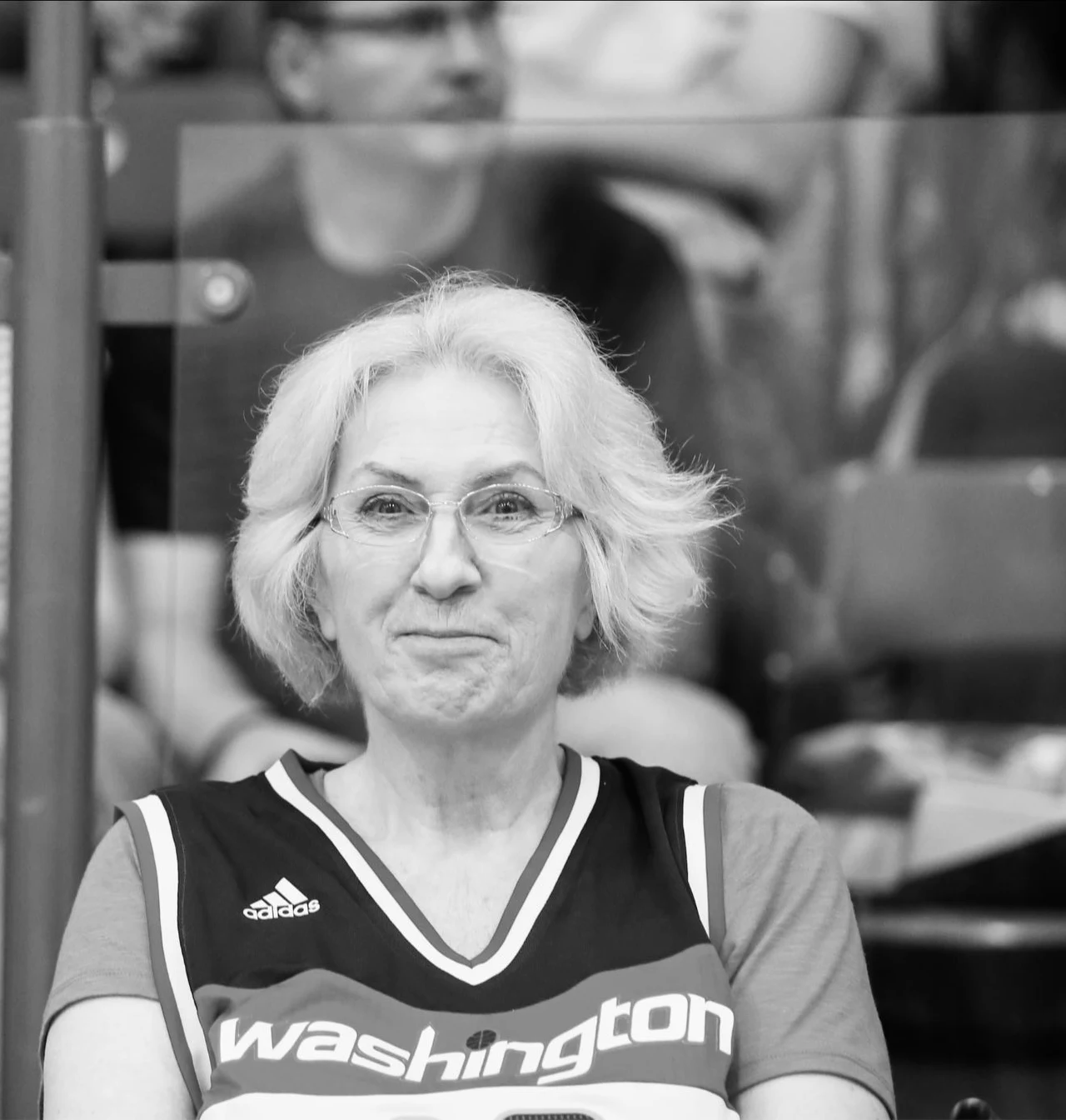 Starsza kobieta z siwymi włosami, w okularach, ubrana w sportową koszulkę z napisem 'Washington', siedzi na trybunach, w tle widoczne rozmyte postacie innych kibiców.