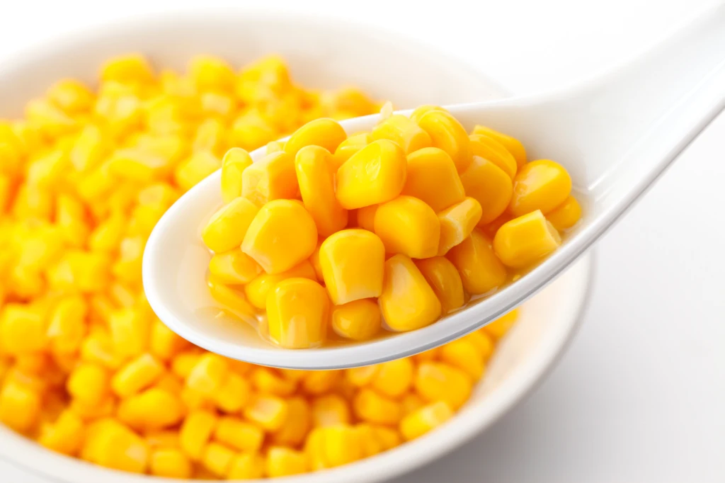 Czy kukurydza konserwowa to dobra alternatywa, kiedy nie mamy dostępu do świeżej kolby? Biała łyżka z ziarnami kukurydzy na tle miski wypełnionej kukurydzą, ziarna są soczyście żółte i błyszczące.