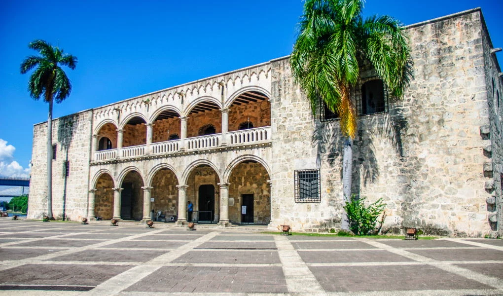 Czas na Twój wymarzony urlop w Dominikanie Historyczny budynek wykonany z jasnego kamienia z arkadami i łukami oraz piętrowym tarasem, otoczony palmami, pod niebieskim niebem.