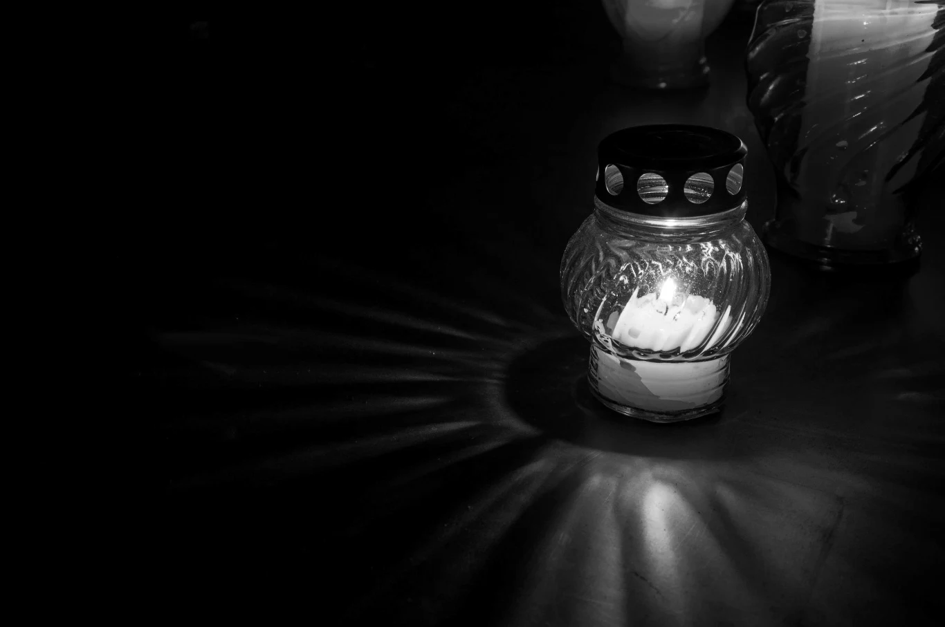 Szklany znicz z zapaloną świecą emitującą delikatne światło, w czarno-białych barwach, z wyraźnie widocznymi promieniami światła odbijającymi się na ciemnym tle. W tle słabo zarysowane kolejne znicze.