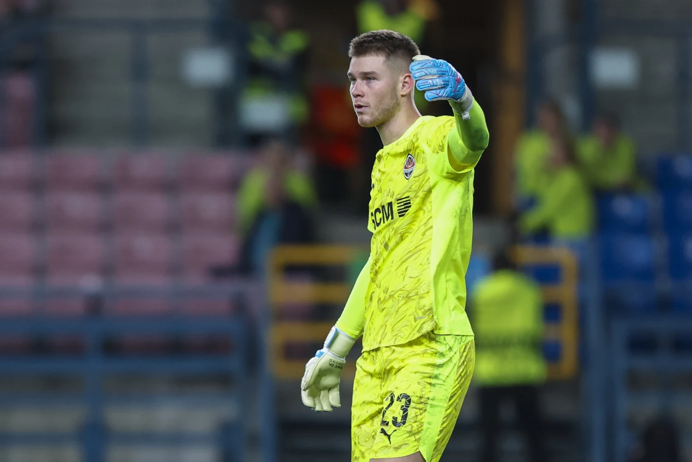 Kirił Fesiun Bramkarz w żółtym stroju bramkarskim, stojący na boisku piłkarskim i wskazujący ręką. Na trybunach widać kilka osób w żółtych kamizelkach odblaskowych.