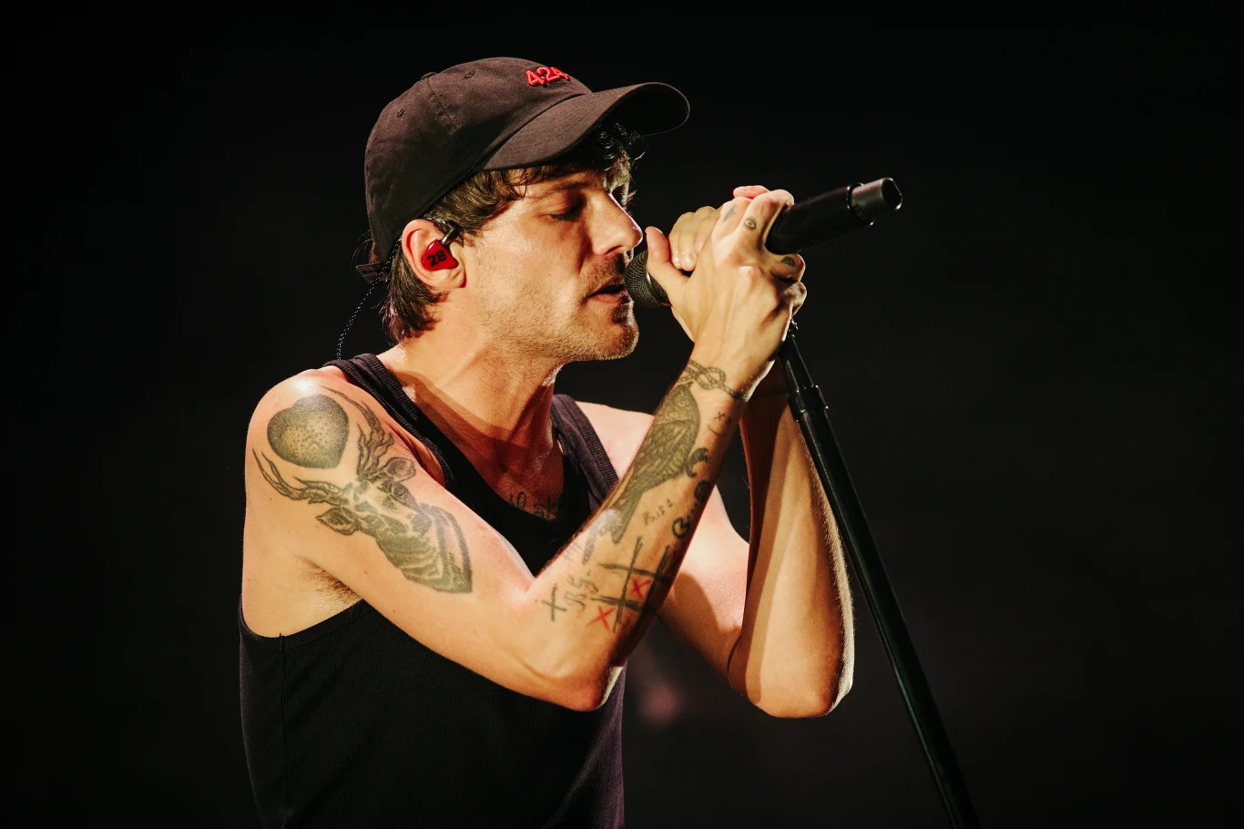 Louis Tomlinson ponownie wystąpi w Polsce Mężczyzna w czarnym podkoszulku i czapce z daszkiem śpiewa do mikrofonu na scenie, z widocznymi tatuażami na ramionach i kolorową słuchawką w uchu, oświetlony światłem scenicznym na ciemnym tle.