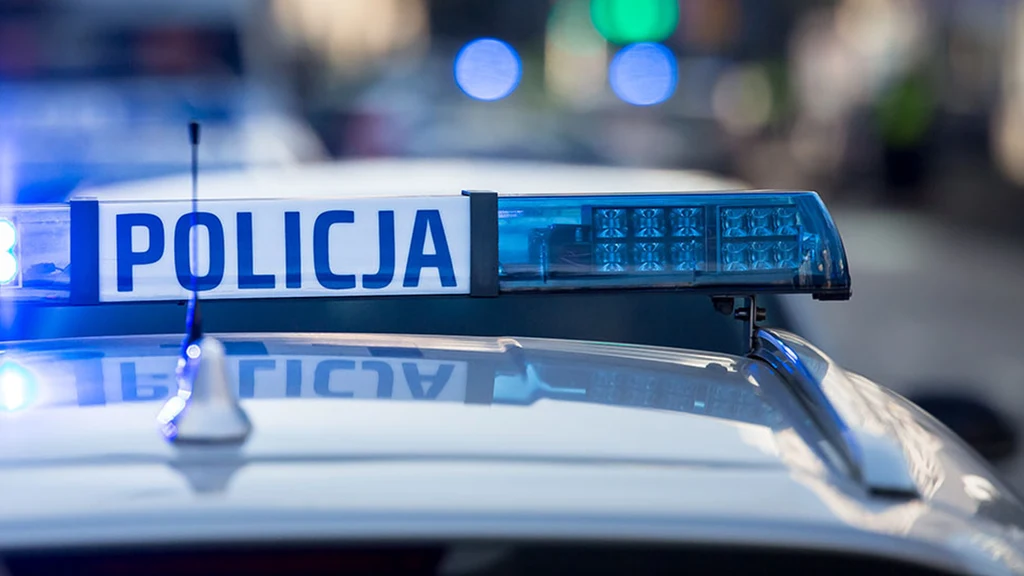 Policja Światła błyskowe niebieskie oraz napis policja na dachu radiowozu policyjnego, rozmyte tło miejskie z widocznym delikatnym ruchem ulicznym.