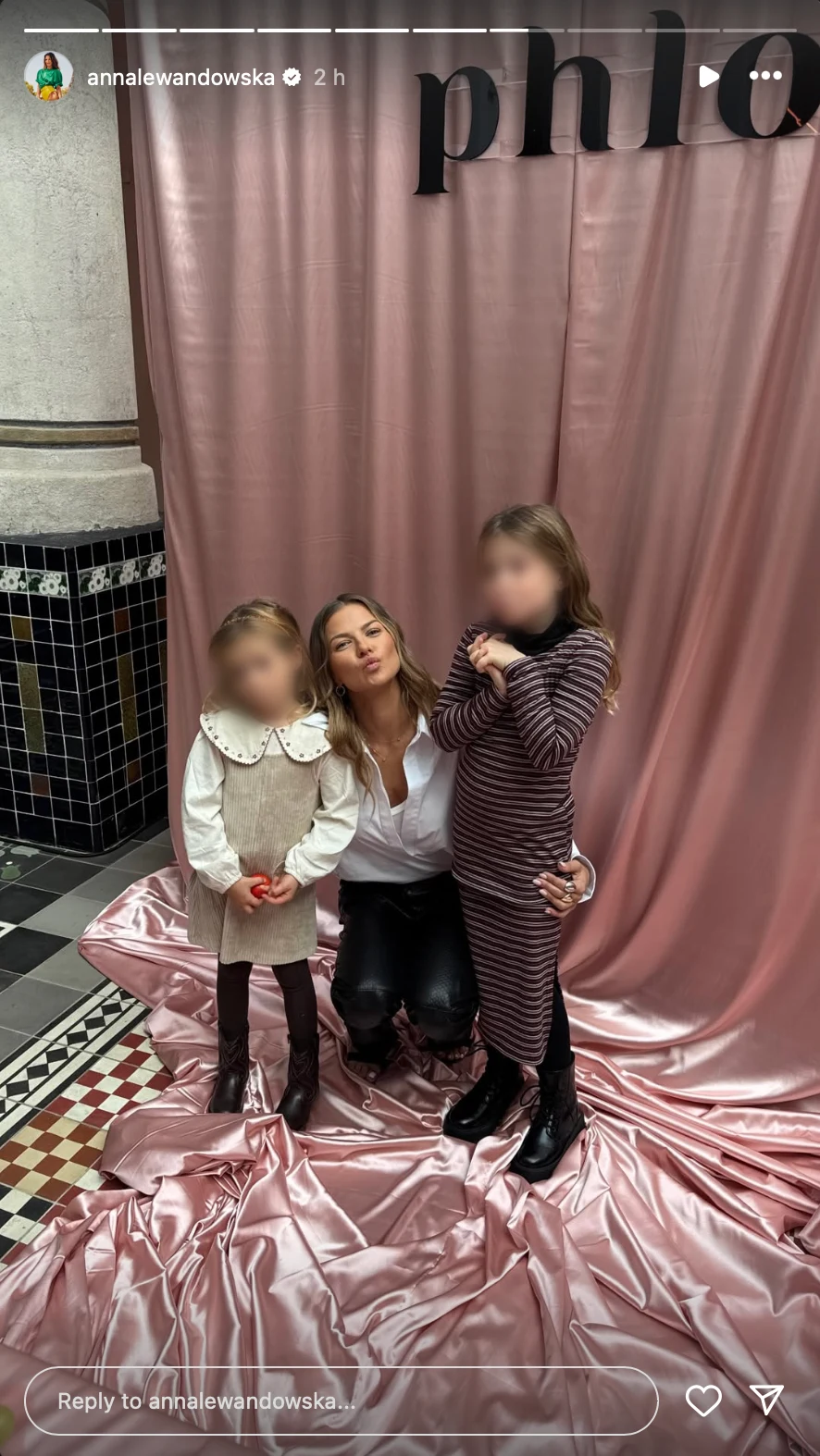 Kobieta klęcząca pomiędzy dwiema dziewczynkami na tle różowej, błyszczącej zasłony i czarno-białej posadzki. Twarze dzieci są rozmyte. Kobieta ubrana w białą koszulę, dziewczynki w jasnej, rozkloszowanej sukience i w prążkowanej sukience z długimi ręka...