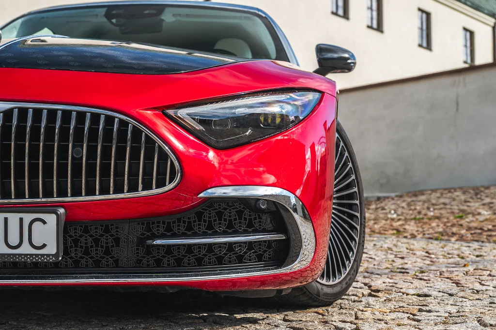 Luksusowe czerwone auto sportowe z charakterystycznym chromowanym grillem, nowoczesnym reflektorem i wypolerowanym nadwoziem zaparkowane na bruku, w tle jasny budynek z oknami.