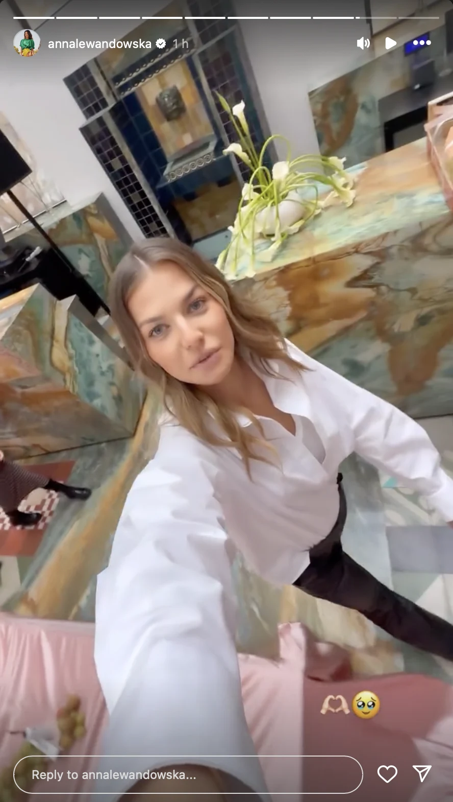 Kobieta w białej koszuli pozuje do selfie w nowoczesnym wnętrzu z marmurowymi blatami i dekoracyjnymi kwiatami w tle.