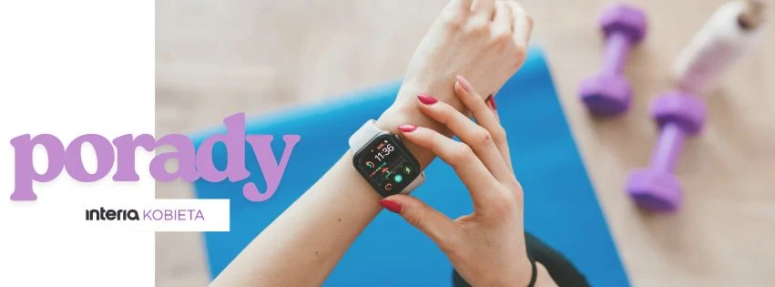 Kobieca ręka z jasno różowym manicure sprawdza dane na smart zegarku, na tle niebieskiej maty oraz dwie małe fioletowe hantle w tle.