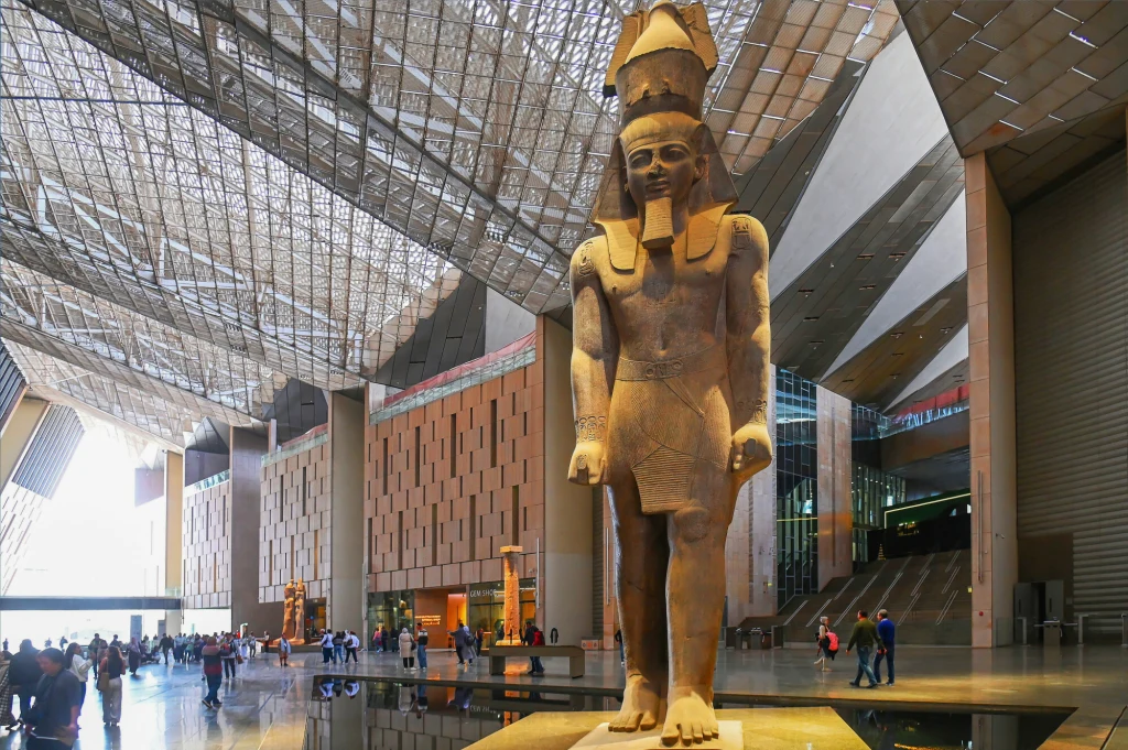 Wnętrze nowoczesnego muzeum z ogromną kamienną rzeźbą faraona na pierwszym planie. Postać stoi na podwyższeniu, wokół spacerują zwiedzający. Nad całością rozpościera się ażurowy, przeszklony dach wpuszczający dużo światła.