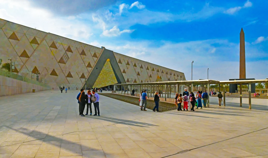 Grupa ludzi spacerująca po przestronnym dziedzińcu przed nowoczesnym budynkiem o geometrycznej fasadzie ze złotymi detalami. Po prawej stronie widoczny obelisk, nad całością rozciąga się błękitne niebo z chmurami.