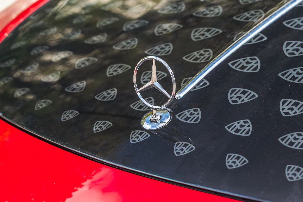 Czerwony samochód z widocznym emblematem Mercedesa na masce oraz licznymi wzorami w kształcie liści lub logo na czarnej powierzchni wokół emblematu.