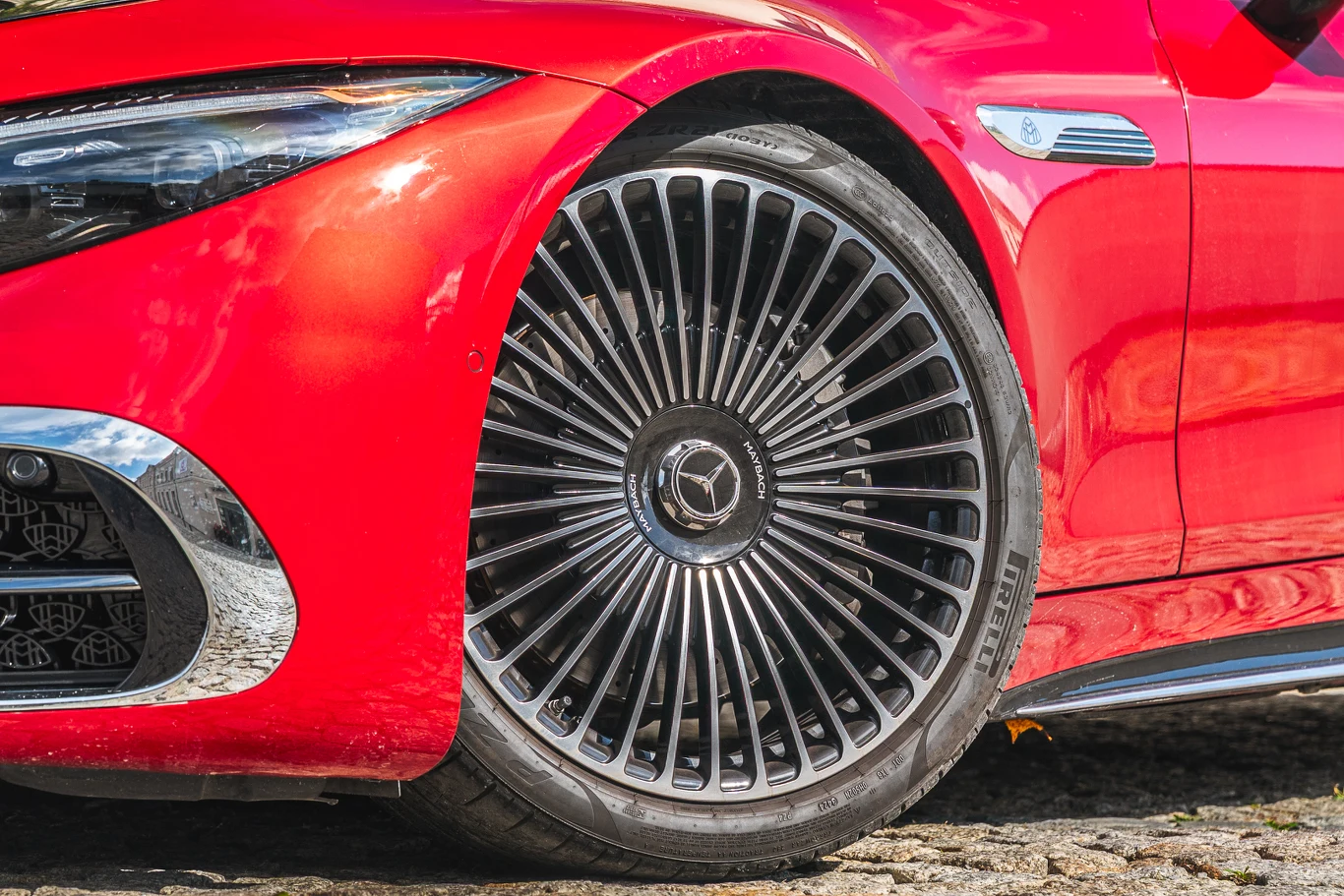 Czerwony samochód marki Mercedes-Benz z widocznym dużym aluminiowym felgiem oraz reflektorem, fragment maski i drzwi, samochód zaparkowany na bruku.