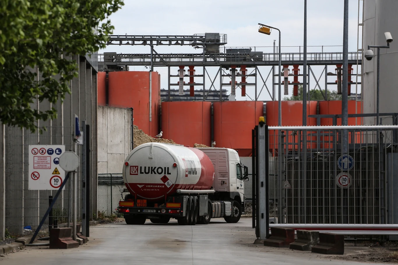 Łukoil sprzeda swoje zagraniczne aktywa grupie Gunvor Cysterna z logo Lukoil wjeżdża przez metalową bramę na teren zakładu przemysłowego, w tle liczne duże, czerwone zbiorniki oraz instalacje przemysłowe, otoczenie ogrodzone siatką i oznaczeniami bezpieczeństwa.