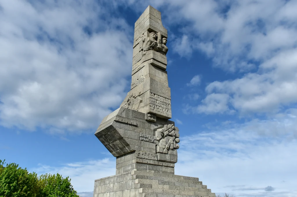 Pomnik Obrońców Wybrzeża na Westerplatte Monumentalna kamienna rzeźba z wyraźnie wyrzeźbionymi twarzami oraz symboliczną dłonią, ustawiona na schodkowej podstawie, otoczona zielenią i na tle nieba z chmurami.