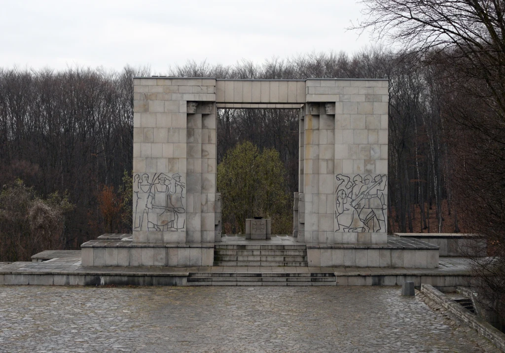 Pomnik Czynu Powstańczego Kamienny monument z geometrycznymi formami oraz płaskorzeźbami po obu stronach, w tle gołe drzewa i szara, pochmurna pogoda.