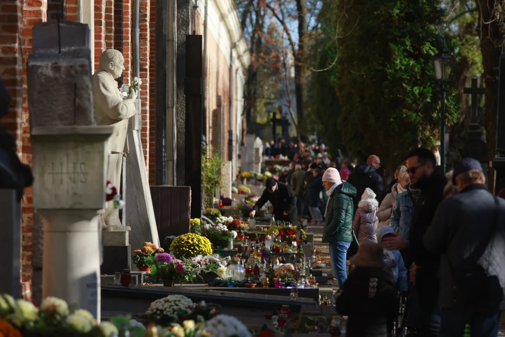 Cmentarz Powązkowski w Warszawie Cmentarz z licznymi nagrobkami udekorowanymi świeżymi kwiatami i zniczami, tłum ludzi odwiedzający groby w słoneczny dzień, po lewej stronie widoczny popiersie świętego lub ważnej postaci religijnej.