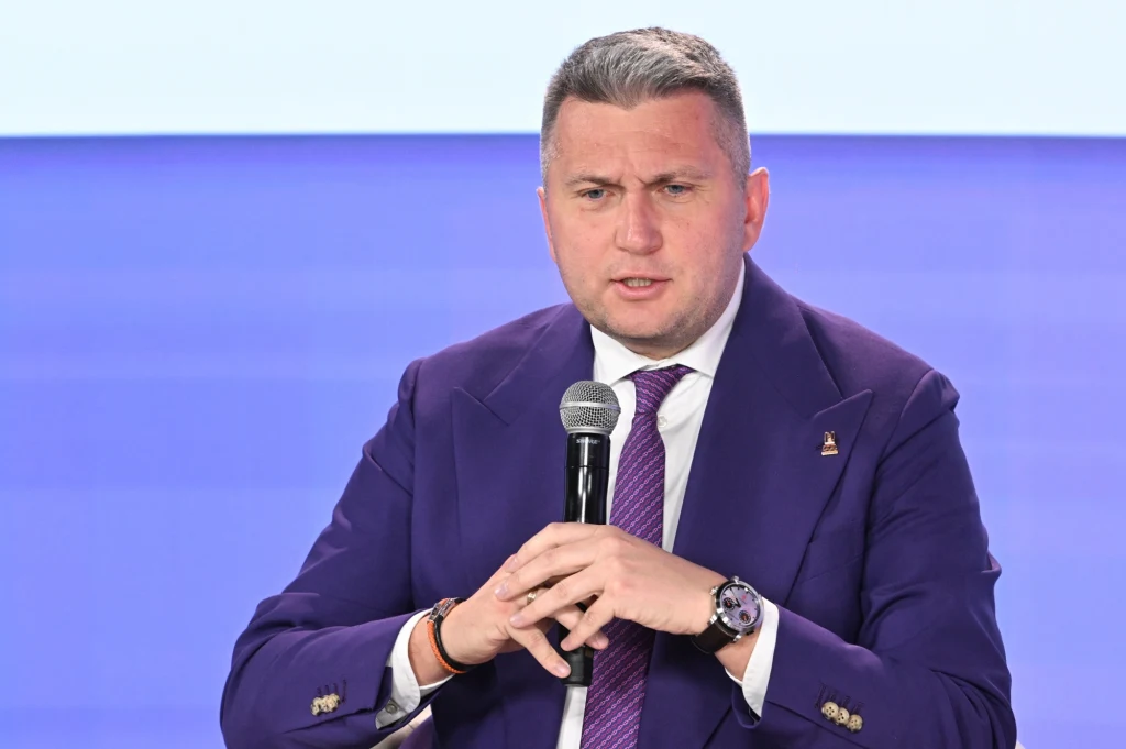 Prezes PKOl Radosław Piesiewicz podczas konferencji prasowej z okazji 100 dni do XXV Zimowych Igrzysk Olimpijskich Mediolan-Cortina 2026 w Lublinie Mężczyzna w ciemnofioletowej marynarce i białej koszuli trzyma mikrofon, siedząc na tle jasnoniebieskiego ekranu. Wygląda na skupionego i zaangażowanego w wypowiedź.
