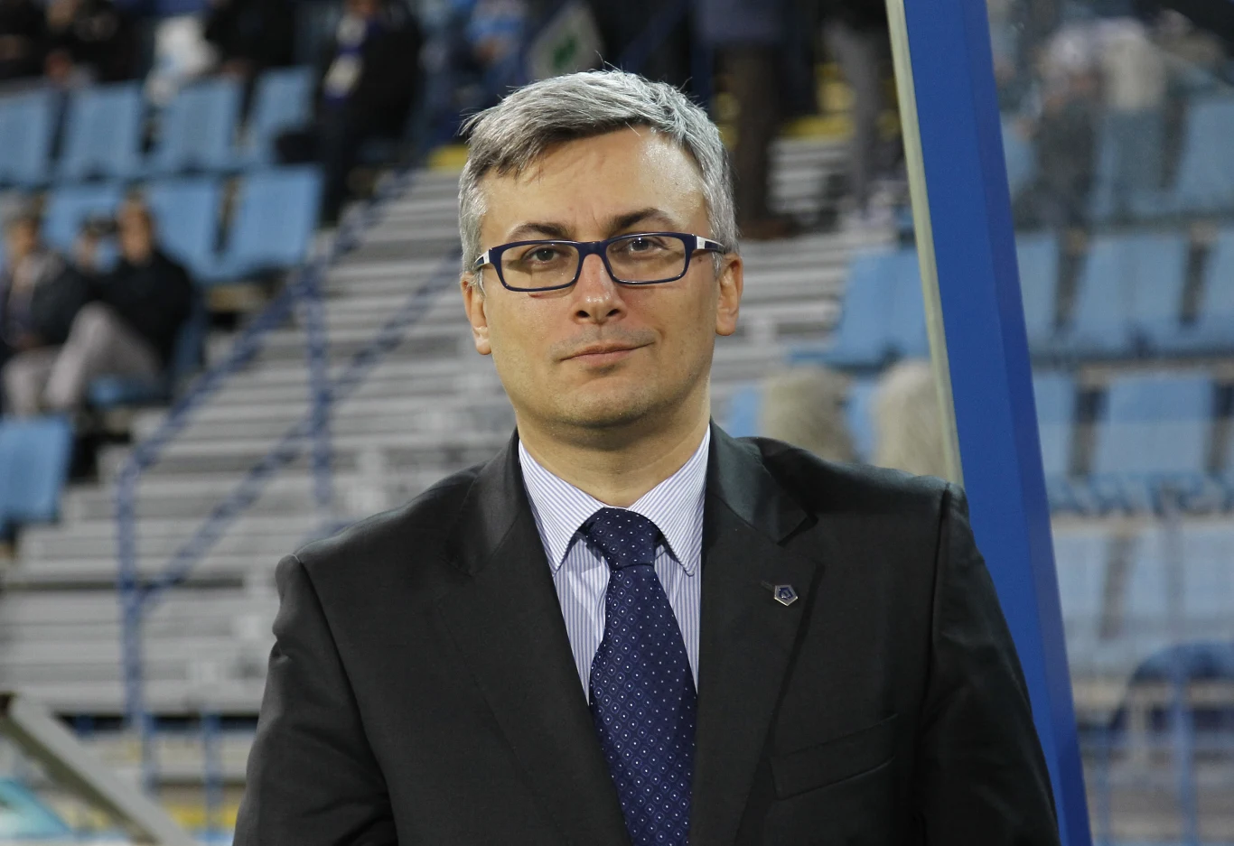 Mężczyzna w okularach i garniturze stoi na tle pustych niebieskich krzeseł stadionowych, z poważnym wyrazem twarzy, w tle widoczne rozmyte sylwetki kilku osób.