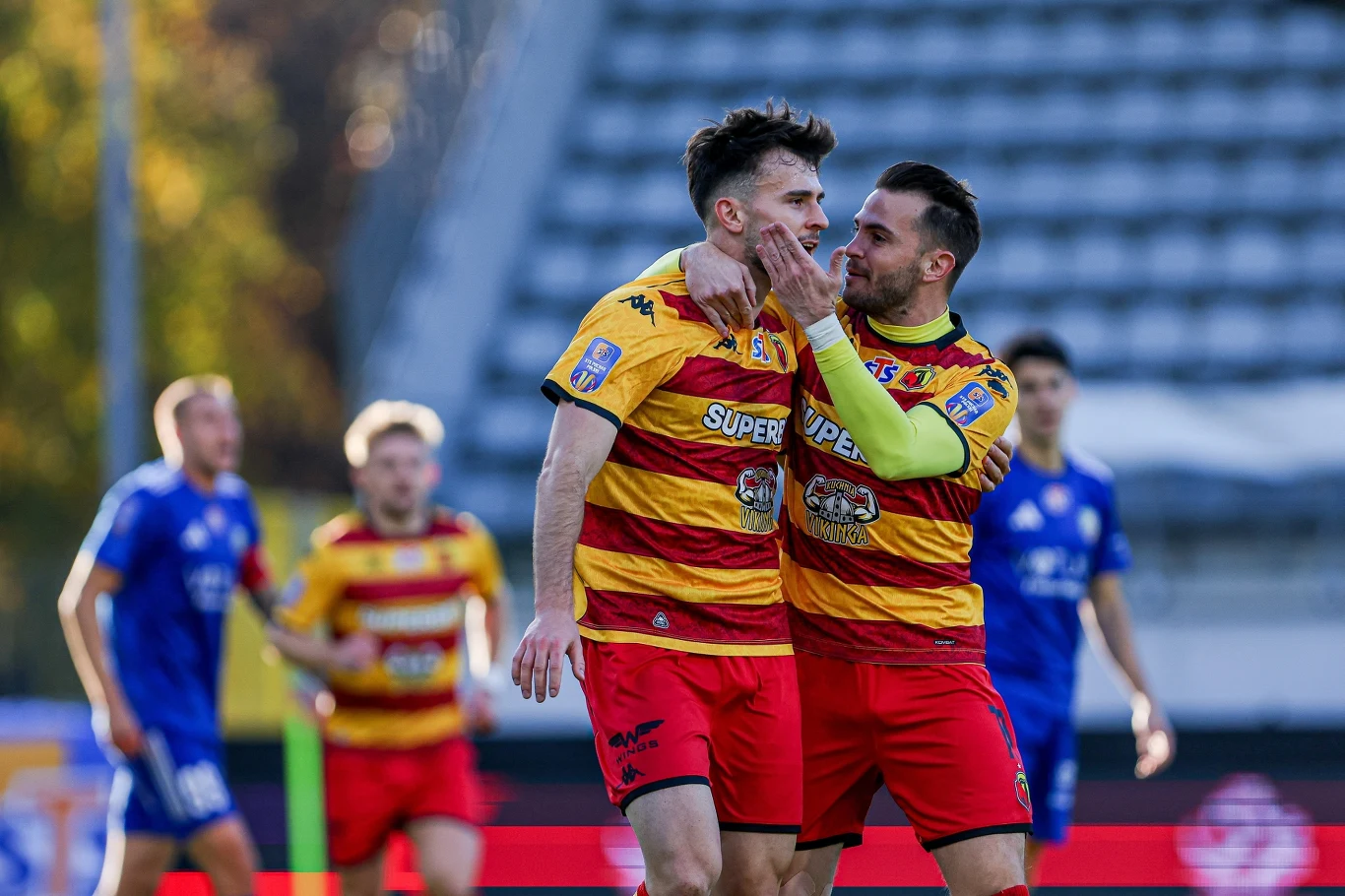 Miedź Legnica - Jagiellonia Białystok w Pucharze Polski Dwóch piłkarzy w żółto-czerwonych koszulkach drużyny świętuje zdobycie gola na boisku, jeden z zawodników obejmuje drugiego, a w tle widoczni są rywale w niebieskich strojach oraz trybuny stadionu.