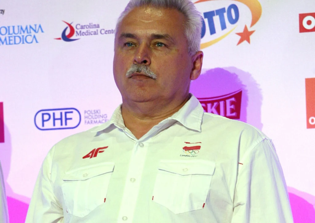 Mężczyzna z siwymi włosami i wąsem, ubrany w białą koszulę z logotypami, stoi na tle ścianki promocyjnej z widocznymi logotypami sponsorów i firm.