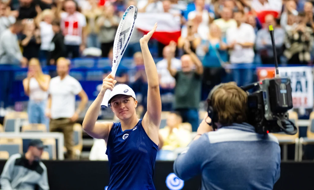 Tenisistka w granatowym stroju i białej czapce unosi rakietę w geście triumfu, znajdując się na korcie podczas zawodów. W tle rozemocjonowany tłum kibiców z polską flagą oraz kamerzysta filmujący wydarzenie.