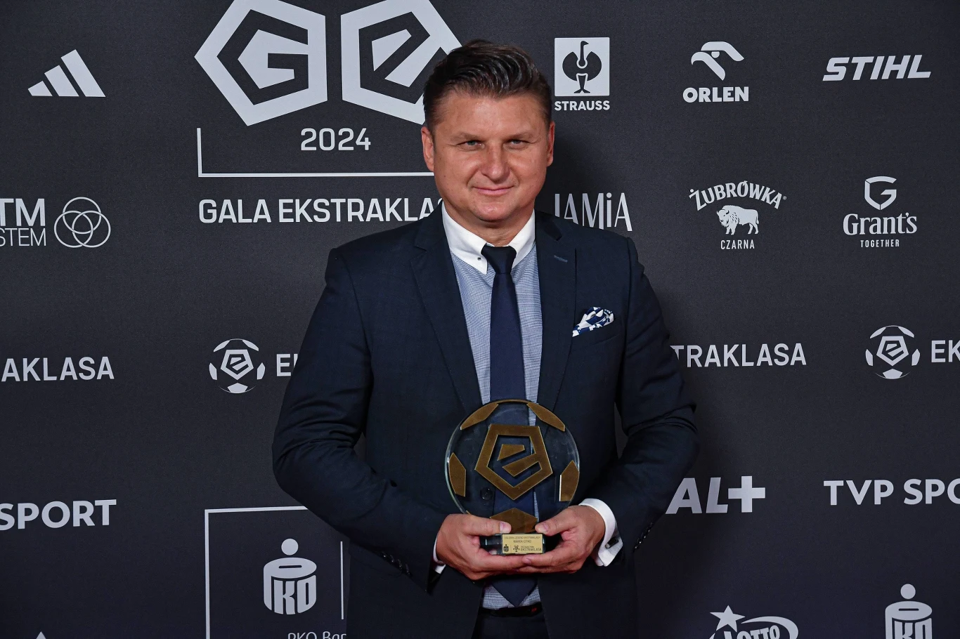 Mężczyzna w eleganckim garniturze trzymający w rękach statuetkę na tle ściany z logotypami sponsorów i organizatorów gali sportowej.