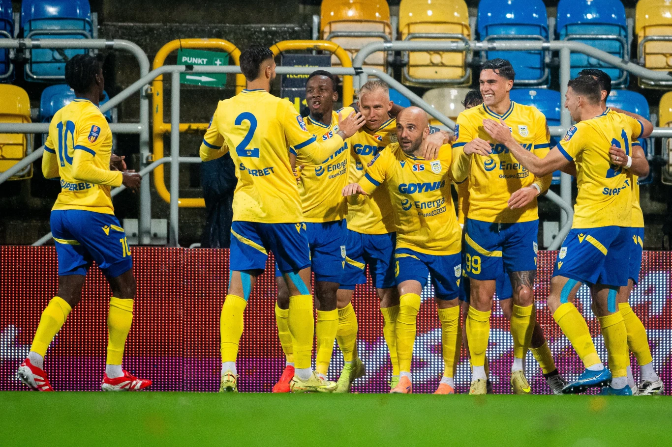 Piłkarze Arki Gdynia Grupa piłkarzy w żółto-niebieskich strojach cieszy się ze zdobycia gola na stadionie, otoczona pustymi, kolorowymi siedzeniami trybun. Zawodnicy okazują emocje, ściskają się i unoszą ręce w geście radości.