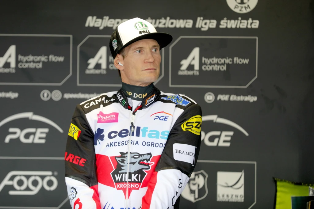 Zawodnik żużlowy w stroju sportowym z licznymi logotypami sponsorów, czapka z daszkiem, w tle tablica sponsora z napisami związanymi z ligą żużlową.