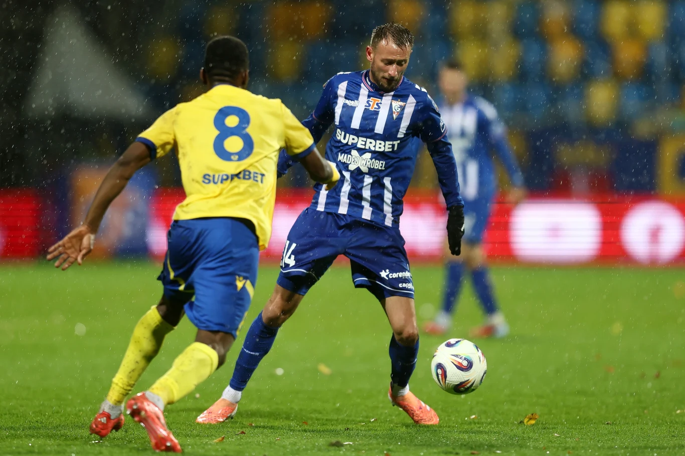 Mecz Arka Gdynia - Górnik Zabrze Dwóch piłkarzy walczy o piłkę podczas meczu piłki nożnej, jeden w żółtej, drugi w niebieskiej koszulce, mokre boisko, w tle rozmyta publiczność i ławka rezerwowych.