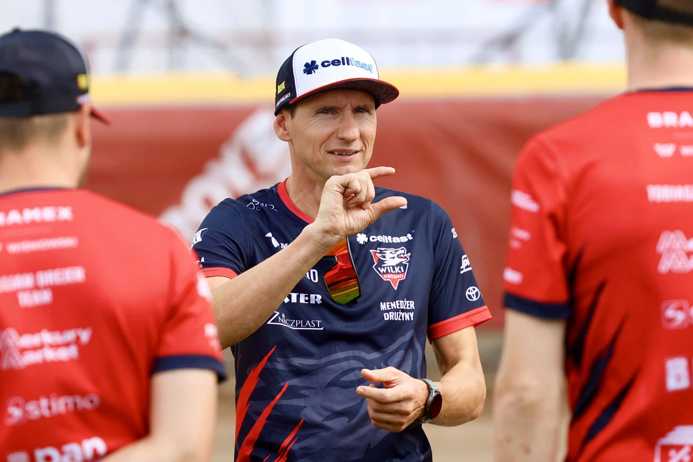 Piotr Świderski, menadżer Wilków Krosno Trzech mężczyzn w czerwono-granatowych koszulkach drużynowych podczas rozmowy na stadionie, jedna osoba w centrum wykonuje gest dłonią, na tle rozmytego boiska.