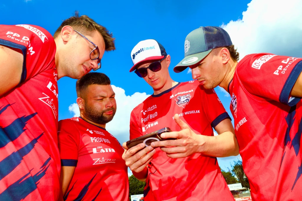 Czterech mężczyzn w czerwonych sportowych koszulkach i czapkach z daszkiem skupia się na wspólnym oglądaniu ekranu telefonu na tle błękitnego nieba z chmurami.