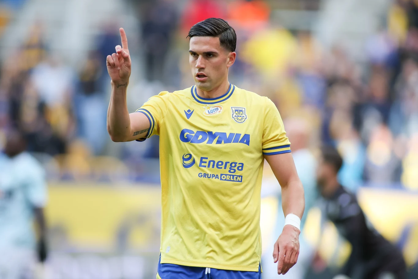 Eduardo David Espiau Hernandez Piłkarz w żółtej koszulce z logo sponsora i nazwą miasta Gdynia unosi rękę z wyprostowanym palcem wskazującym w górę. Na tle zamazani pozostali zawodnicy oraz trybuny stadionu.